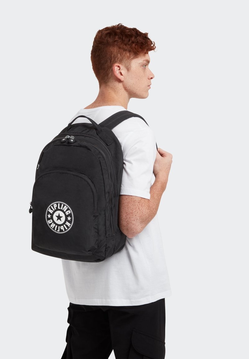 Mochila de nylon negra con forma redondeada, que cuenta con dobles cremalleras y un logotipo circular en la parte frontal. Las correas ajustables mejoran la comodidad.