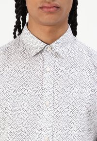 Camicia bianca con bottoni, decorata con un piccolo motivo navy, colletto a punta e due bottoni visibili nella parte superiore. Il tessuto appare liscio e leggero.
