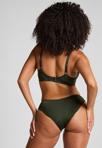 Femme aux cheveux bouclés portant un soutien-gorge vert foncé et une lingerie assortie, posée de manière à montrer son dos et les mains reposant sur ses hanches.