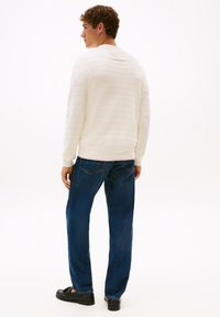 Weiß gestreifter Pullover, blaue Jeans und schwarze Schuhe. Der Pullover hat einen runden Ausschnitt und gerippte Bündchen. Die Jeans haben einen klassischen Schnitt.