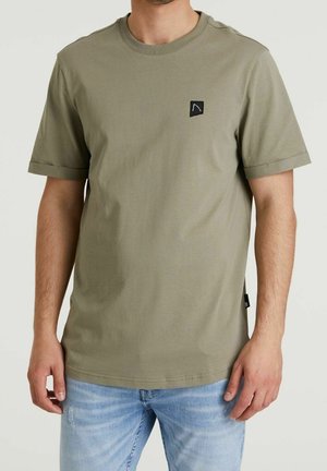 Mężczyzna ubrany w oliwkowozielony t-shirt z krótkim rękawem z małym czarnym logo na klatce piersiowej oraz jasnoniebieskie, wyblakłe jeansy, stojący na jednolitym tle.