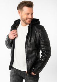 Giubbotto di pelle nera con cappuccio rimovibile, chiusura con zip e tasche laterali; indossato sopra una camicia bianca e jeans grigi. Finitura texturizzata.