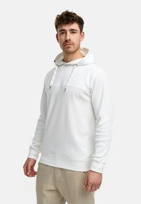 Witte hoodie met textuurstof, contrasterend glad paneel en koorden. Gecombineerd met lichte beige broek.