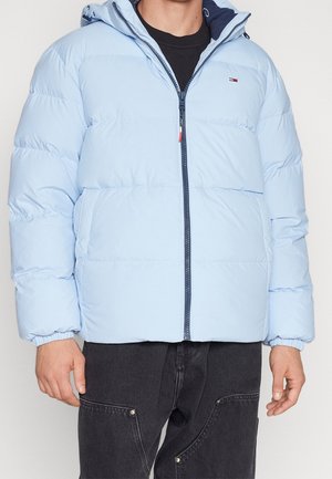 Muž ve světle modré péřové bundě na zip s elastickými manžetami a černých kalhotách s velkými předními kapsami.