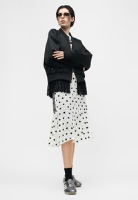 Άτομο που φοράει γυαλιά ηλίου, μαύρο μπουφάν τύπου bomber πάνω από midi φόρεμα με λευκές πουά, μαύρες κάλτσες και ασημί αθλητικά παπούτσια, στέκεται με σταυρωμένα τα χέρια.