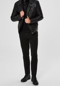 Man i svart läderbikerjacka, svarta byxor, svart turtleneck och svarta snörstövlar, stående mot en enfärgad bakgrund.