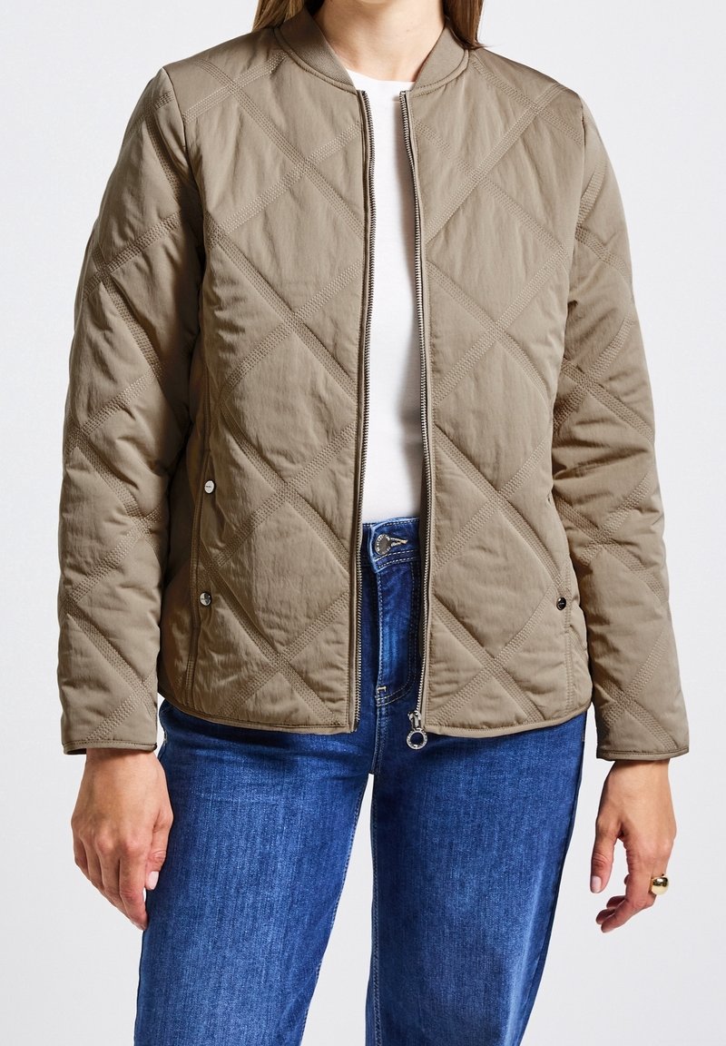 Femme portant une veste matelassée marron clair zippée par-dessus une chemise blanche et un jean bleu, debout les bras le long du corps.