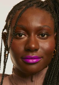 Gros plan sur une personne à la peau foncée et aux cheveux tressés, portant un rouge à lèvres violet vif, des boucles d'oreilles en or et un haut zippé marron clair.