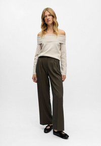 Beige schulterfreier Pullover, kombiniert mit dunkelgrünen weiten Hosen und schwarzen flachen Schuhen. Glattes Material mit geripptem Ausschnitt.