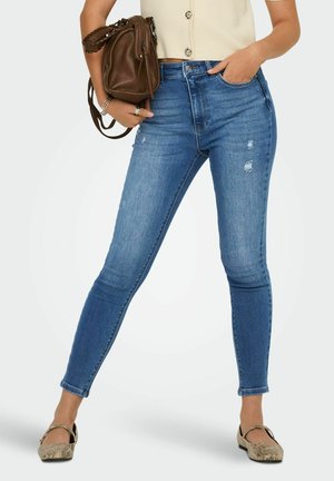 Kobieta w niebieskich obcisłych jeansach z wysokim stanem z lekkim postarzaniem, beżowym krótki topem, trzymająca brązową torebkę i nosząca płaskie buty ze wzorem wężowej skóry.