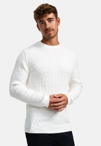 Hvit kabelstrikket genser med rund hals, teksturert mønster og ribbestrikkede mansjetter og kant. Stoffet ser mykt og koselig ut.