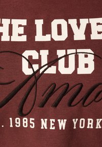 Merlotgekleurde sweatshirts met witte blokletters waarop staat "THE LOVE CLUB" en een zwarte geborduurde tekst "Mama" in sierlijke letters, met daaronder "1985 NEW YORK".