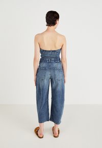 Jeansoverall med axelbandslös, smockad överdel, vida byxor, blekblå färg och två bakfickor. Bärs med bruna slip-on sandaler.