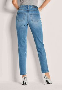 Eine Frau trägt hellblaue, gerade geschnittene Jeans und silberne High Heels. Sie steht mit den Händen in die Hüften gestützt vor einer einfarbigen Wand.