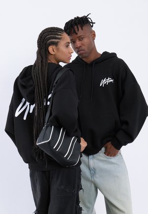 Deux jeunes adultes portant des sweat-shirts noirs avec des inscriptions blanches, l'un tenant un sac noir à rayures blanches, debout près l'un de l'autre sur un fond blanc.