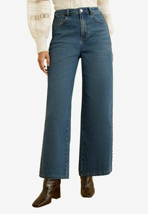 Žena má na sebe modré rifle s vysokým pásom a širokými nohavicami, hnedé čižmy na podpätku a bielu blúzku s dlhým rukávom s čipkovými detailmi.