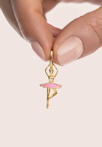 Złota zawieszka w kształcie baleriny z różową spódniczką tutu, z misternymi detalami na figurce i pętelką do zawieszenia, trzymana między palcami.
