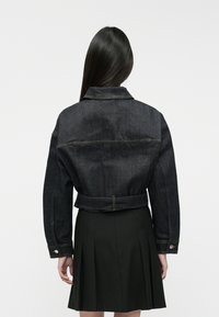 Chaqueta de mezclilla negra con un diseño corto, costuras visibles y cintura con cinturón; llevada sobre una falda negra con pliegues.