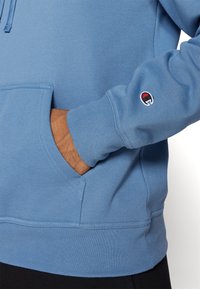 Svijetloplava dukserica s kenguruzim džepom, rebrastim manžetama i pojasom. Sadrži mali izvezeni logotip na lijevom rukavu. Materijal je mješavina pamuka.