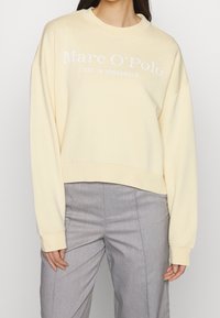 Sweat-shirt jaune clair avec un col rond et des épaules tombantes. Il présente un texte blanc indiquant "Marc O'Polo EST. À STOCKHOLM" sur le devant.