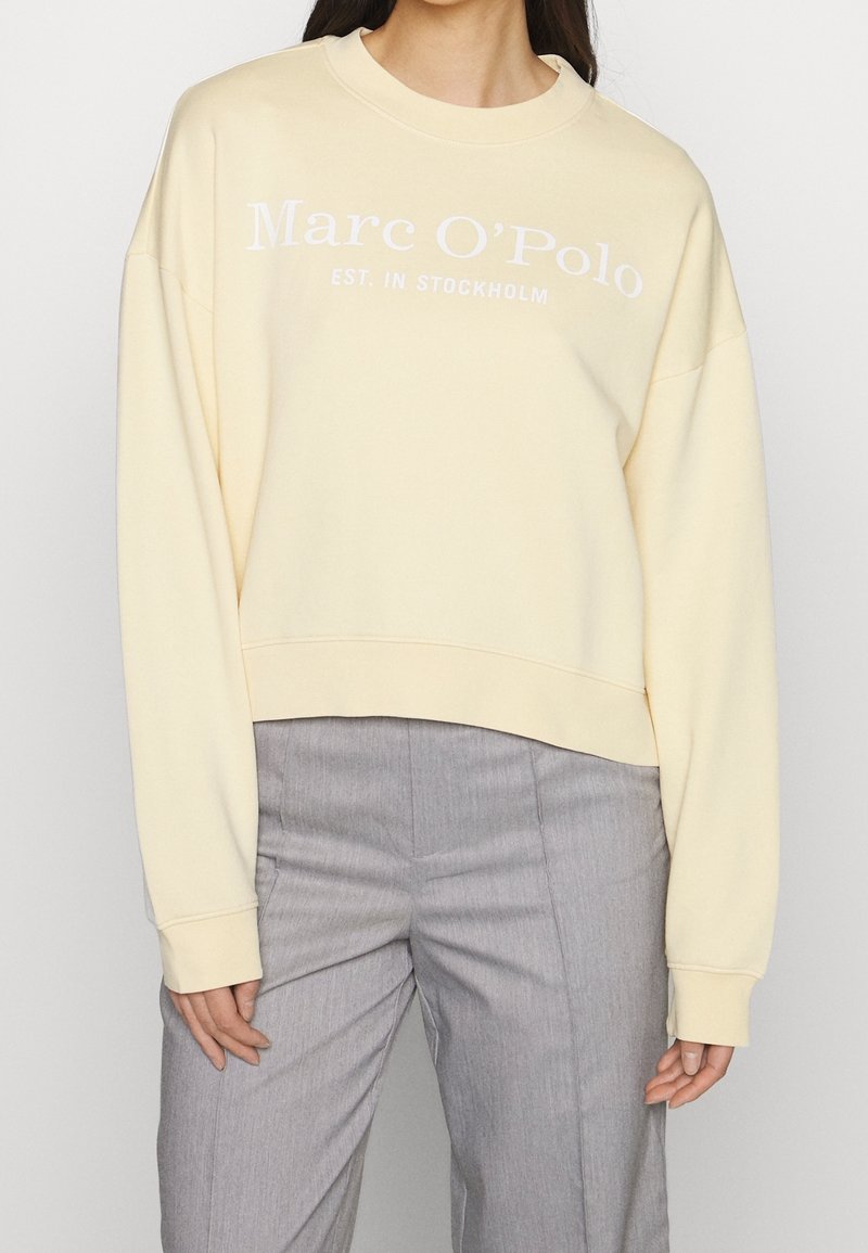 Sweat-shirt jaune clair avec un col rond et des épaules tombantes. Il présente un texte blanc indiquant "Marc O'Polo EST. À STOCKHOLM" sur le devant.