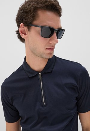 Uomo con occhiali da sole neri e polo blu navy con zip che guarda di lato su uno sfondo uniforme.