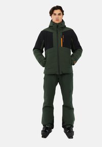 Giacca isolante verde con accenti neri, tasca sul petto con zip, e colletto alto, abbinata a pantaloni verdi coordinati e scarponi da sci.