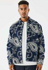 Giacca di denim con uno sfondo blu navy e complessi ricami in paisley bianchi, chiusura a bottoni e vestibilità rilassata. Indossata sopra una t-shirt bianca.