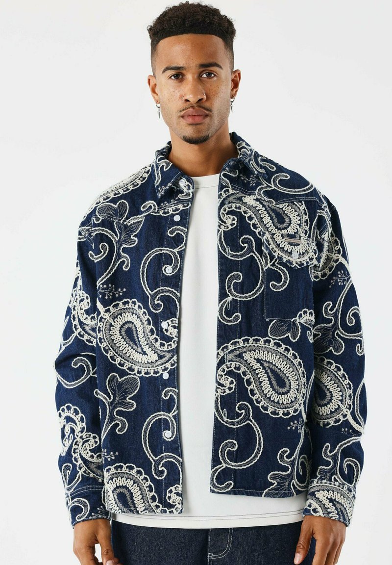 Giacca di denim con uno sfondo blu navy e complessi ricami in paisley bianchi, chiusura a bottoni e vestibilità rilassata. Indossata sopra una t-shirt bianca.