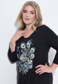 Schwarzes langärmliges Oberteil mit einem Pfau-Graphic in Blau und Creme. Weicher Stoff mit einem Rundhalsausschnitt und lockerer Passform.