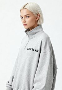 Grijze sweater met rits, met een hoge kraag en een losse pasvorm, met zwarte geborduurde tekst "C'est la vie" aan de voorkant. Zachte textuur.
