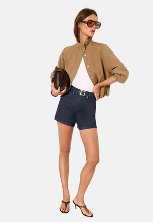 Naine kannab üle suurendatud päikeseprille, päevitunud lühemate varrukatega jopet valge topi peal, tumedaid kõrge vöökohaga lühikesi pükse, hoiab käes pruuni klambrikotti ja pruune kontsakingi.