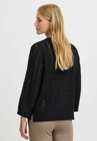 Pull en tricot noir avec un motif de mailles ouvertes, des poignets et un ourlet côtelés. Coupe ample, épaules tombantes et fentes latérales pour faciliter le mouvement.