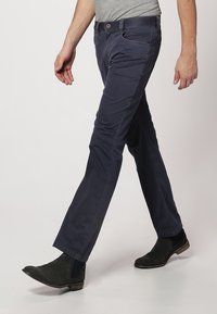 Mężczyzna idący, ubrany w granatowe spodnie, szarą koszulkę i czarne botki na gładkim tle.