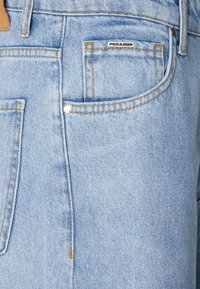 Jeans in denim azzurro chiaro con tasca dotata di bottone a pressione argentato, cuciture gialle e una piccola etichetta "PEGANOR" sulla tasca superiore.