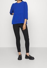Blusa blu a maniche lunghe con spalle arricciate, vita peplum, indossata con pantaloni neri skinny e scarpe slip-on nere chunky.