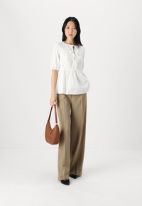 Bílý peplum top s výřezem, doplněný vysokým béžovým kalhotami s širokými nohavicemi. Modelka drží hnědou kabelku.