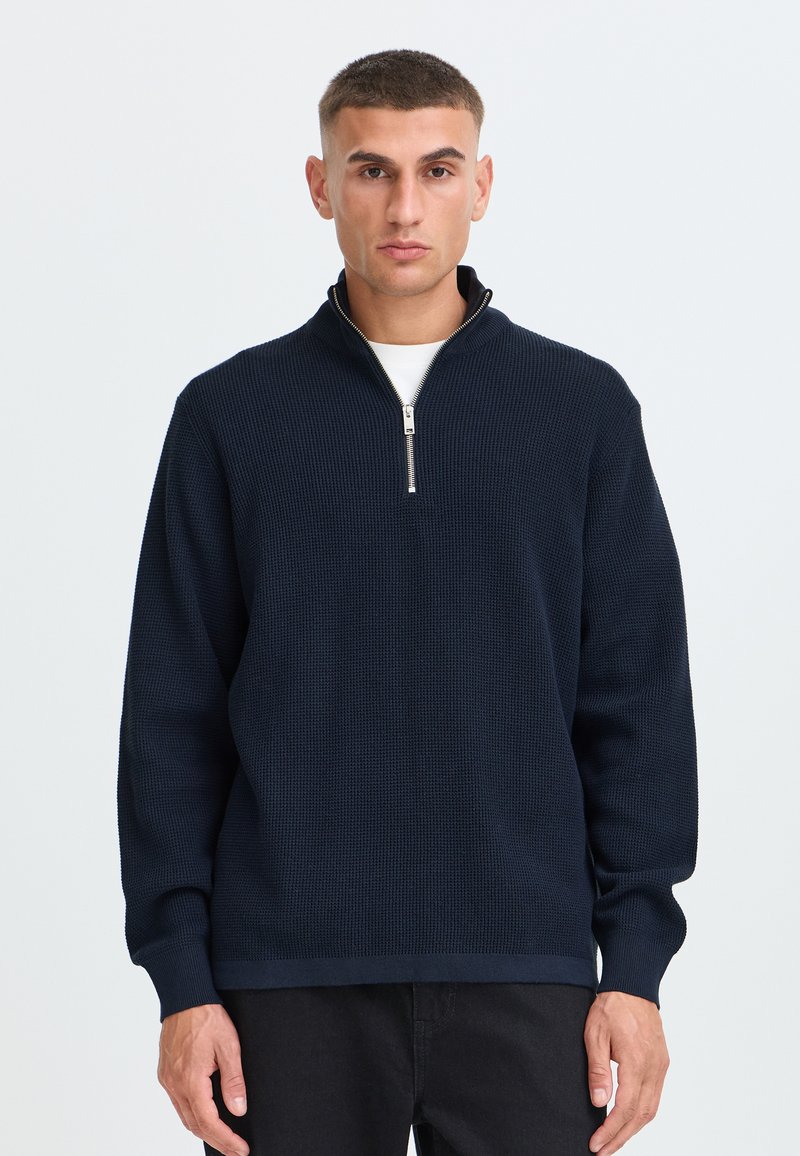 Homme aux cheveux courts portant un pull en maille bleu foncé avec une fermeture éclair à moitié ouverte par-dessus une chemise blanche, debout devant un fond clair uni.