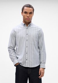 Pelēkā svītraina pogājamā krekls ar apkakli, garām piedurknēm, vienu krūšu kabatu un plastmasas pogām. Valkāts ar tumšiem denima džinsiem.