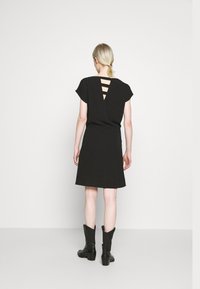 Mujer vistiendo un vestido negro de manga corta con cortes triangulares en la parte posterior y botas negras hasta la mitad de la pantorrilla, de pie frente a una pared blanca.