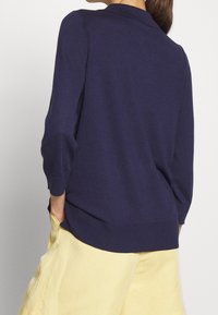 Mörk marinblå stickad tröja med en mjuk textur, trekvartslånga ärmar och krage. Kombineras med ljusgula vida byxor.