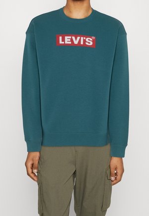 Person iført en blågrøn crewneck-sweatshirt med rød og hvid Levi's-logo i midten af brystet og olivengrønne cargobukser.