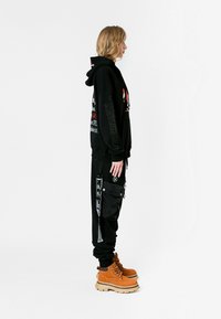 Persona con sudadera gráfica negra y pantalones cargo con parches, llevando botas de cordones color beige, de pie de lado contra un fondo blanco.