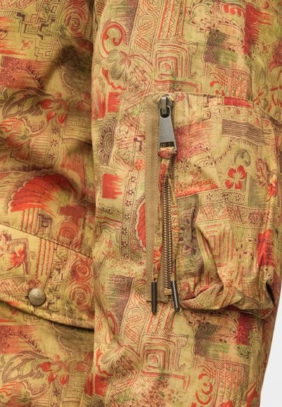 Vzory na látke v tlmených zelených, červených a béžových odtieňoch. Obsahuje vrecko na zips s kovovým kovaním a nariasené rukávy pre textúru.