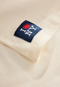 Krém színű pamut póló, amelyen egy kék négyzet alakú folt látható, fehér és piros "I ♥ NY" felirattal. Az anyag puhasága és a látható varrások jól kivehetők.