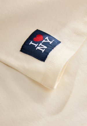 Kreemivärvi puuvillane T-särk, mille keskel on sinine ruudukujuline plaastriga, millel on valge ja punasega kiri "I ♥ NY". Kangas näib pehme ning õmblused on nähtavad.