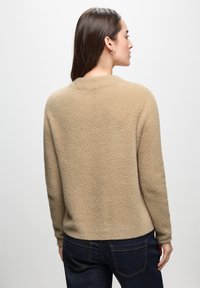 Beige, fluffig stickad tröja med rundad halsringning och långa ärmar, kombinerad med mörkblå jeans. Mjuk textur och avslappnad passform.