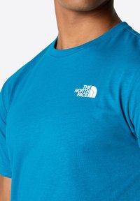Camiseta de algodón azul con cuello redondo; presenta un logo blanco de "The North Face" en la parte izquierda del pecho. Textura suave.