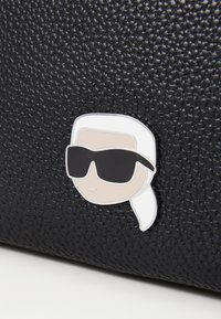 Borsa nera con texture che presenta un volto di cartone incorniciato con capelli bianchi e occhiali da sole neri come dettaglio decorativo.