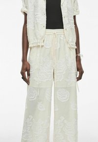 Chaqueta corta blanca bordada con pantalones de pierna ancha a juego, que presentan grandes patrones florales y una cinturilla elástica con cordón.
